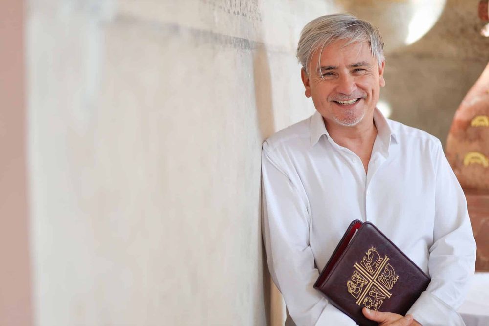 Udo Quass lehnt lachend an einer hellen Wand und hält ein Buch