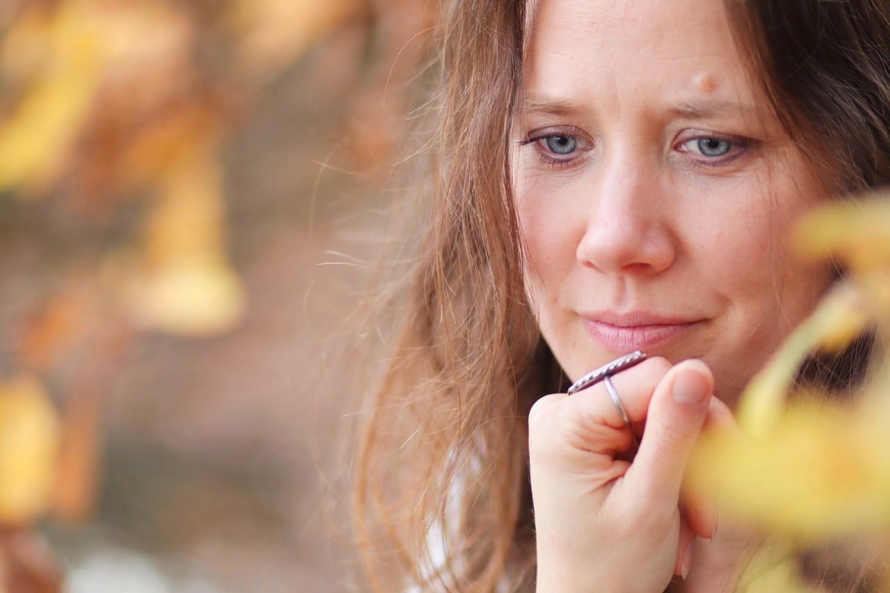 Nohila schaut nach unten, die Hand am Kinn, vorn gelbe Blätter